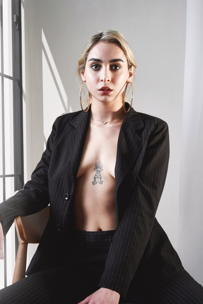 Fernanda Padilla a model with a striking appearance poses in a minimalist setting. She wears a pinstriped blazer revealing a tattoo on her chest and large hoop earrings. The lighting casts subtle shadows emphasizing her facial features and the contours of her body.